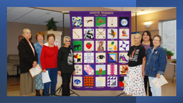 Photo of seven indigenous women standing by a quilt